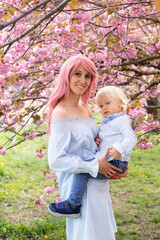 Young woman with a child in her arms happy. Sakura blossom in spring Park. Joy of motherhood and child development. Cherry blossom tree in late spring