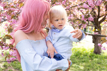 young mother and little son are having fun in the blooming Sakura gardens. Portrait woman hugging little baby boy