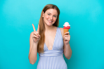 Young caucasian woman in swimsuit eating ice cream isolated on blue background smiling and showing...