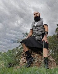 low angle image of a very masculine mature man with black beard dressed in skirt leaning on a palm stump with cloudy sky behind him © anuatmoralesfoto