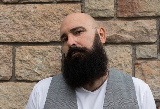 close up portrait of a head-shaved man with black beard standing ahead a stone brick wall