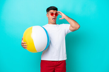 Fototapeta premium Young caucasian man holding a beach ball isolated on blue background doing surprise gesture while looking to the side