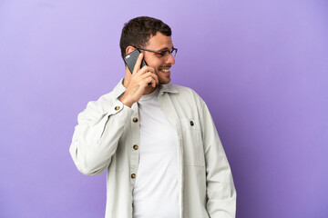 Brazilian man over isolated purple background keeping a conversation with the mobile phone with someone