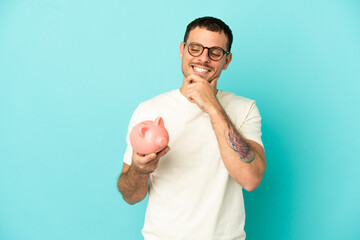 Brazilian man holding a piggybank over isolated blue background looking to the side and smiling