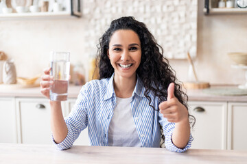 Excited latin woman holding glass with clean water and showing thumb up, sitting at table in...