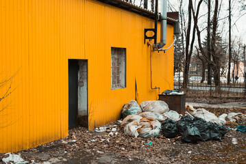 Abandoned industrial building, unfavorable area. Side view of yellow wall, door, barred window and pile of garbage in bags