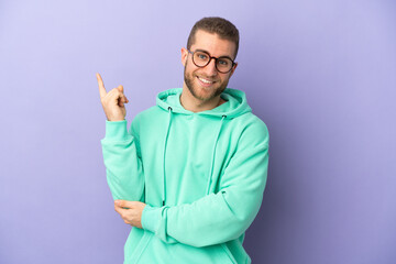 Young handsome caucasian man isolated on purple background happy and pointing up