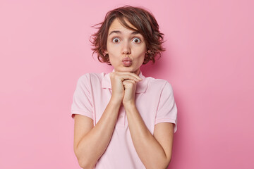 Fototapeta premium Stunned pleasant looking woman with short hair keeps hands under chin lips folded has widely opened eyes dressed in casual t shirt isolated over pink background. Human facial expressions concept