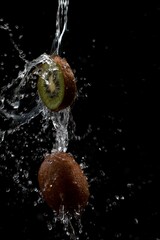 a slice of kiwi and a whole kiwi are doused with plenty of water in front of a black background