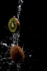 a slice of kiwi and a whole kiwi are doused with plenty of water in front of a black background