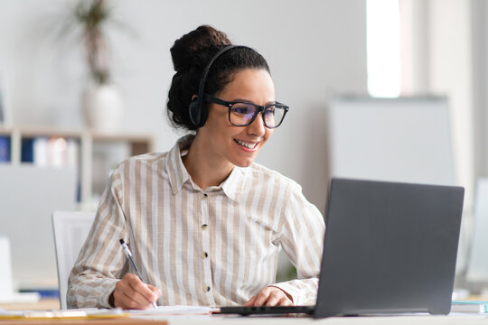 Portrait of happy young latin woman wearing wireless headphones watching online course, sitting at table