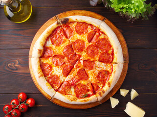 Traditional Italian pepperoni pizza with salami and cheese on a dark brown wooden background. Top view with space to copy. Olive oil, cherry, lettuce. Menu of cafes, pizzerias, restaurants. Close up.