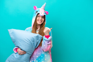 Young caucasian girl with unicorn pajamas holding pillow isolated on blue background pointing to the side to present a product