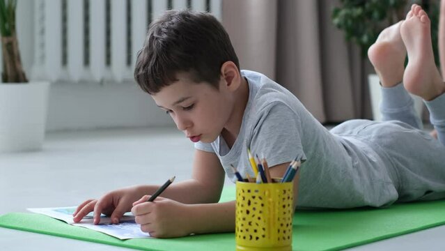 School boy lying on floor and drawing with pencil. cute kid, boy making notes in diary at sunny day. child plays at home on the warm floor.