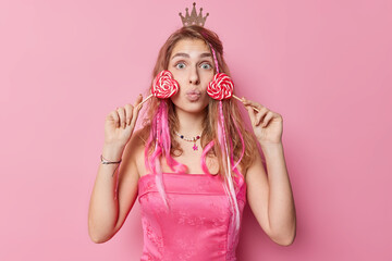 Obraz na płótnie Canvas Horizontal shot of surprised young woman holds heart shaped lollipops near face keeps lips folded wears dress and small crown on head isolated over pink studio background. Sweet tooth concept