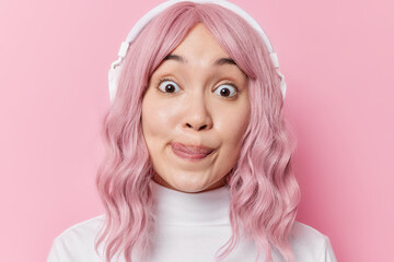 Surprised beautiful young Asian woman has healthy skin widely opened eyes licks lips with tongue dressed in casual white turtleneck listens music via headphones poses against pink background