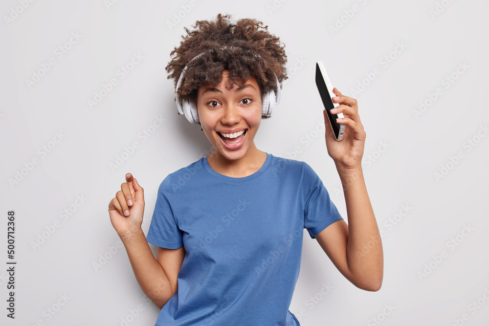 Wall mural Positive curly haired adorable woman dances to music holds mobile phone listens favorite song via wireless headphones wears casual blue t shirt isolated over white background feels energetic