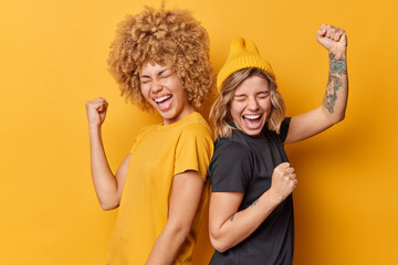 Fototapeta Two positive women friends feel very happy like winners make triumph gesture shake arms celebrate success stand back to each other dressed in casual t shirts isolated over yellow background. obraz