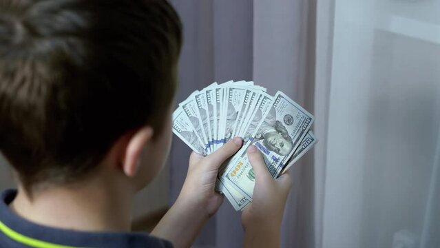 Successful Teenager Counts a Stack of 100-Dollar Bills in Room. Back view. Pack of banknotes in hands. A rich boy learns how to make money. Triumph. Earnings. Wages, profit. Finance, wealth, capital.
