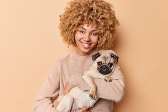 Friendship concept. Smiling beautiful young woman carries her pug dog spends free time with favorite pet wears casual jumper isolated over brown background. Domestic animal with its caring owner