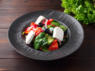 Fresh Greek salad with tomatoes, cucumbers, bell peppers, olives, purple onions and feta cheese on a black plate, top view, dark wooden background. Vegan salad with tofu cheese. Copy space. Close up.