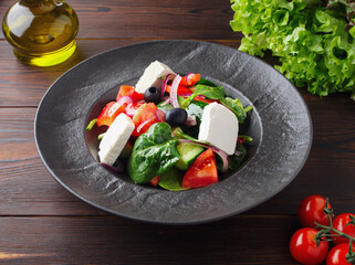 Fresh Greek salad with tomatoes, cucumbers, bell peppers, olives, purple onions and feta cheese on a black plate, top view, dark wooden background. Vegan salad with tofu cheese. Copy space. Close up.