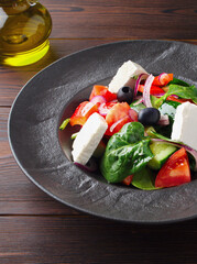 Fresh Greek salad with tomatoes, cucumbers, bell peppers, olives, purple onions and feta cheese on a black plate, top view, dark wooden background. Vegan salad with tofu cheese. Copy space. Close up.