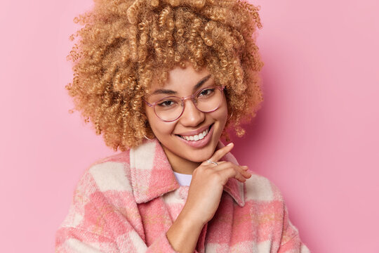 Portrait of lovely woman has natural curly hair touches jawline gently  smiles toothily looks happily at camera dressed in fashionable coat  isolated over pink background. Pleasant feelings concept Stock Photo | Adobe