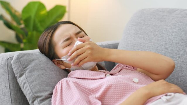 Young Asian woman infected with coronavirus covid-19 have a cough and sore throat during home isolation. Lying down on sofa and wearing mask. Pandemic of disease