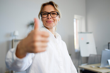 Happy middle aged business woman showing thumb up while standing in office