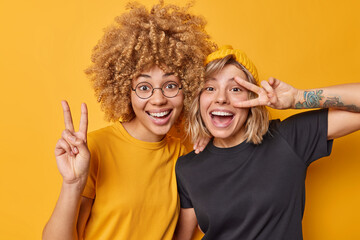 Positive two female friends show peace gesture smile gladfully laugh happily dressed in casual t shirts isolated over yellow background. Playful girls go crazy have good mood make victory sign