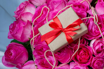 A gift with a red satin ribbon lies on a bouquet of pink roses with a string of pearls