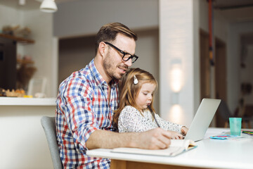 Young father using computer while his little girl sitting on his lap at home