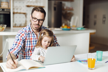 Young father using computer while his little girl sitting on his lap at home