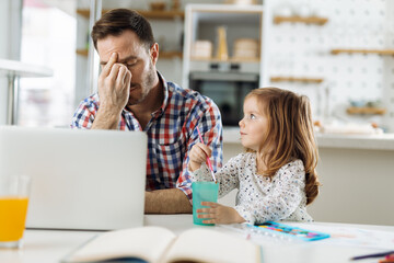 Tired father working on laptop from home while his little girl is sitting next to him