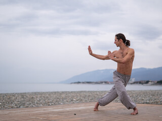 Caucasian man with naked torso practicing wushu on the seashore. 