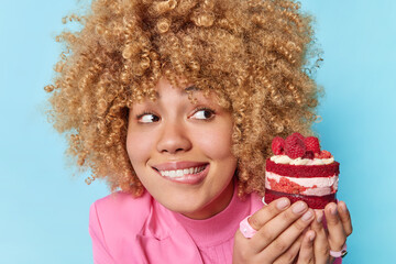 Curly haired beautiful woman bites lips looks at appetizing piece of cake with raspberries has...