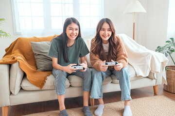 Two happy asian roommates sitting on couch in living room at home enjoy and excited holding console...