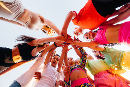 A large group of children, boys and girls, stand together in a circle and fold their hands, tuning up and raising team spirit before the game at the stadium during sunset. A healthy lifestyle. - Powered by Adobe