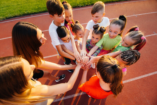 A large group of children, boys and girls, stand together in a circle and fold their hands, tuning up and raising team spirit before the game at the stadium during sunset. A healthy lifestyle.
