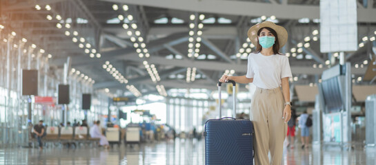 Fototapeta premium Young female wearing face mask with luggage walking in airport, protection Coronavirus disease infection, Asian woman traveler with hat. Time to travel after vaccine booster dose concept