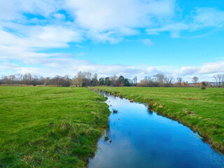 A glassy stream reflects blue sky as it meanders through a wide-open expanse of water meadow from a tree lined horizon.