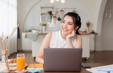 Beautiful young asian woman working on laptop computer while sitting at the kitchen room background.