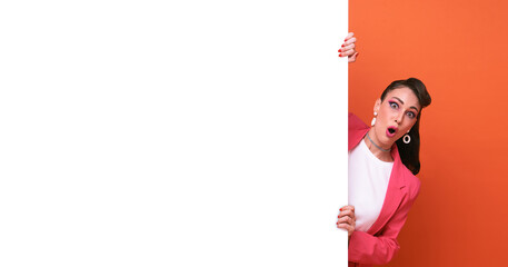 Surprised woman in pink blazer with hand holding blank announcement banner in orange background.