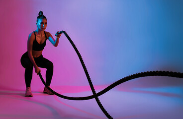 Athlete asian sportswoman waving the ropes as part of fat burning workout in fitness studio neon background. Woman exercising with battling ropes at the gym.