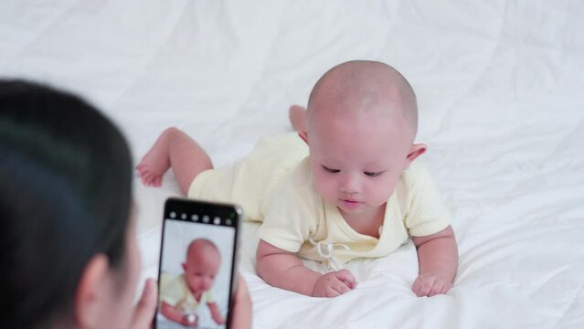 Happy family, Mother taking photo or video, cute newborn with mobile phone. Asian baby play enjoy on white bed at home. Little innocent new infant adorable. Technology, lifestyle parenthood concept.