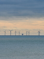 The silhouetted turbines of a wind farm, guarded by the hulk of a Maunsell Fort, stand on the horizon in the North Sea under a dramatic, stormy sky.