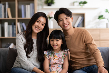 Happy Asian family sitting on couch smile with love and looking at camera spend time together.Father mother and little daughter relax on sofa in cozy living room.Good moment.Happy and warmth family.