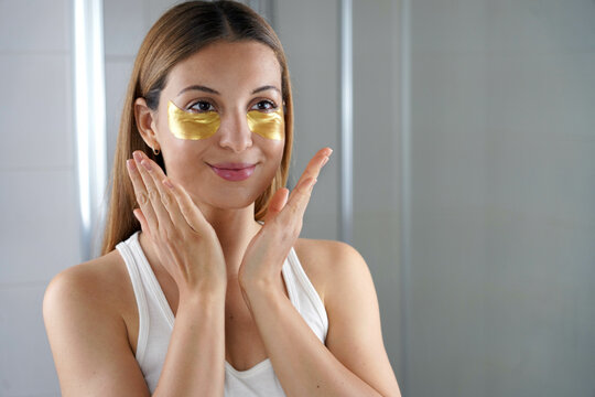 Beauty Girl Applying Golden Anti-aging Under-eye Mask Looking Herself In The Mirror In The Bathroom. Skin Care Woman With Patches Of Fabric Mask Under Eyes To Reduce Eye Bags.