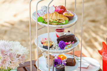 Afternoon tea on tropical sandy beach with shadows from palm trees in hot summer day. Cake stand with fresh pastries, sandwiches, sweeties, shake. Travel in Thailand, luxury hotel and restaurant